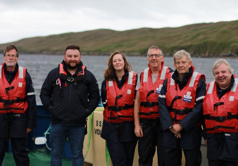 Six people wearing life jackets stand in front of a water body with hills behind them.