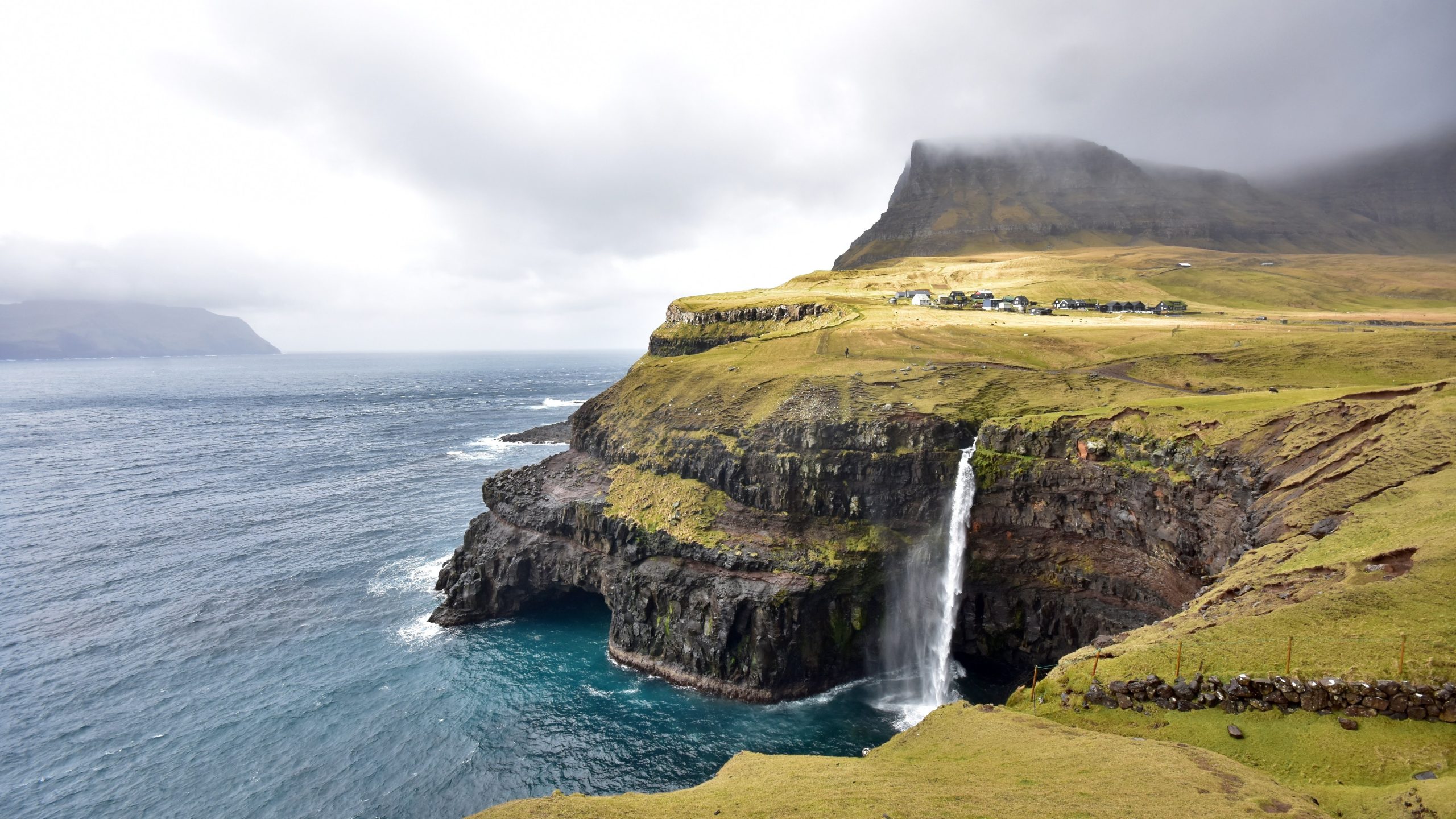 Richard McCulloch Gasadalur, Faore Island Spectacular view of Gásadalur waterfall cascading from a grassy cliff into the Atlantic Ocean on the Faroe Islands, with rugged mountains and a small village under dramatic cloudy skies.