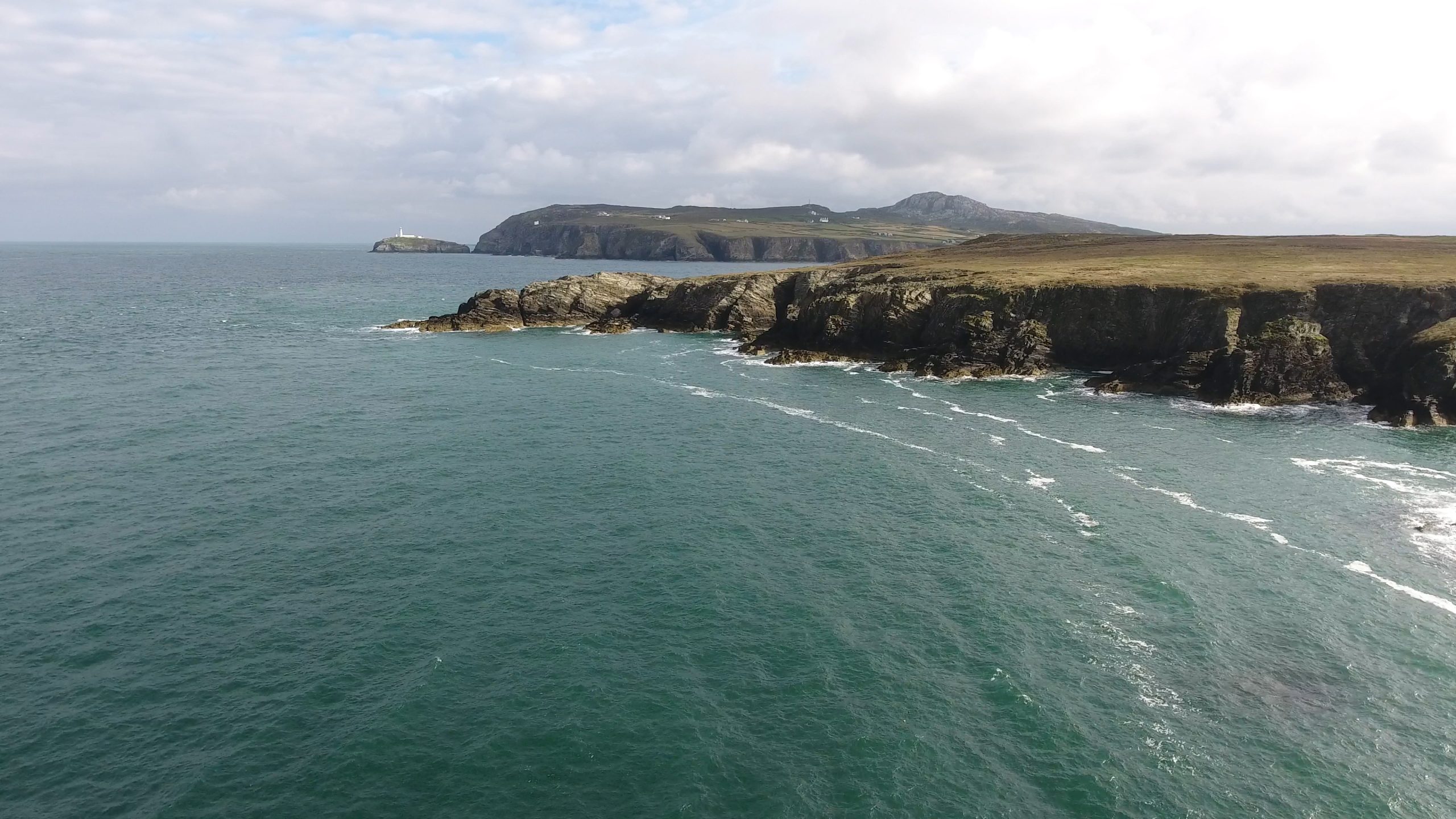 Aerial view of rugged coastal cliffs and deep blue sea at RSPB nature reserve on Anglesey, Wales, with a distant lighthouse and rolling hills under a partly cloudy sky.