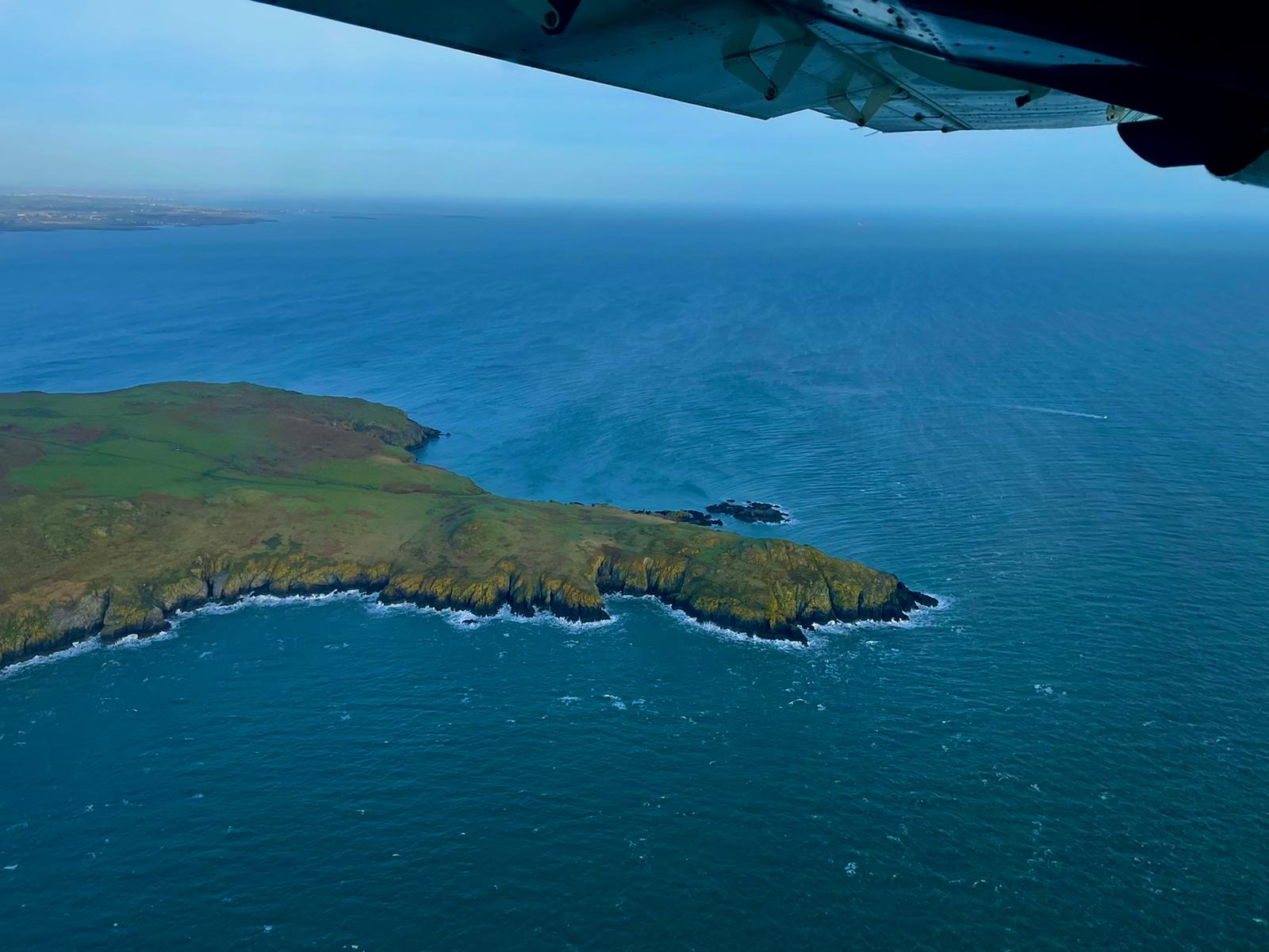Aerial view of a rugged green coastline in Ireland surrounded by deep blue sea, with part of an aircraft wing visible overhead.